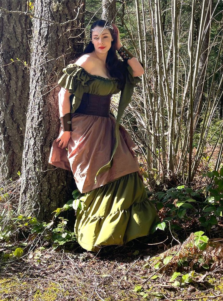 a woman in a green and brown dress standing next to a tree with her hands on her head