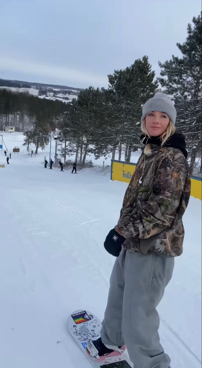 a young person on a snowboard standing in the snow with trees and people behind them