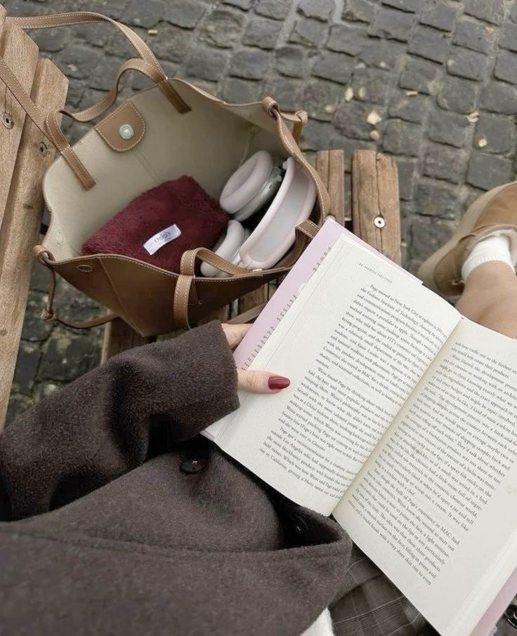 a person sitting on a bench with a book in their lap and purse next to them