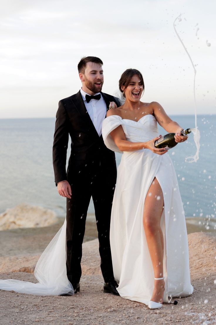 a newly married couple standing next to each other near the ocean with champagne in their hands