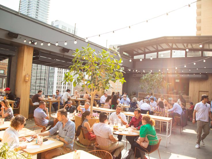 many people are sitting at tables and having drinks in an outdoor dining area with string lights strung across the roof