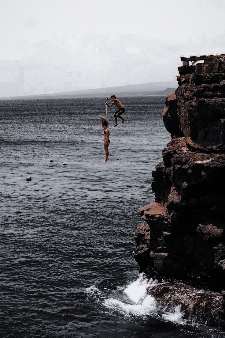 two people jumping off rocks into the ocean from a rocky cliff near an ocean shore