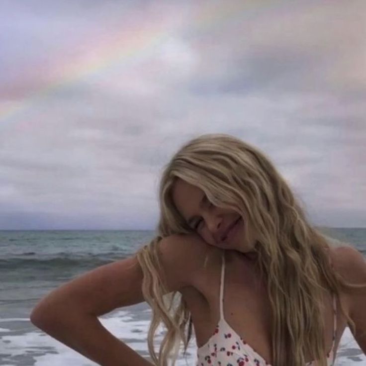 a woman standing on top of a beach next to the ocean with a rainbow in the sky