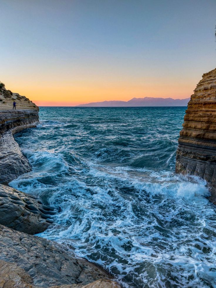 the sun is setting over the ocean with waves crashing on the rocks and people standing at the edge