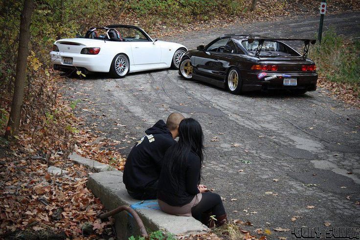 two people are sitting on the curb next to their cars