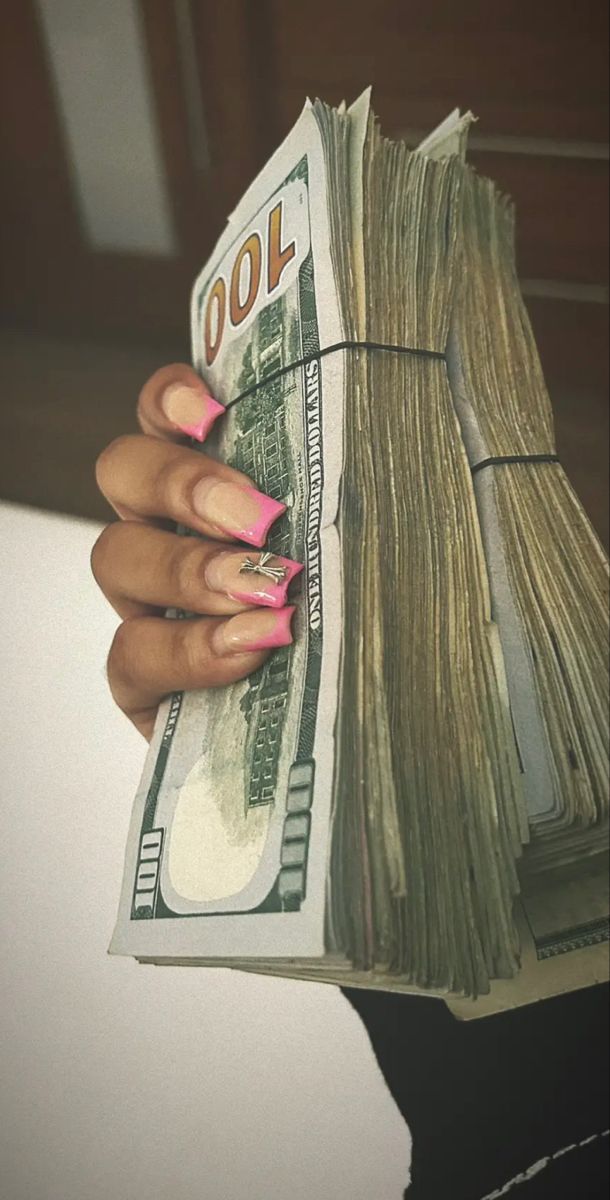 a woman's hand holding a stack of money in front of her face with pink nails