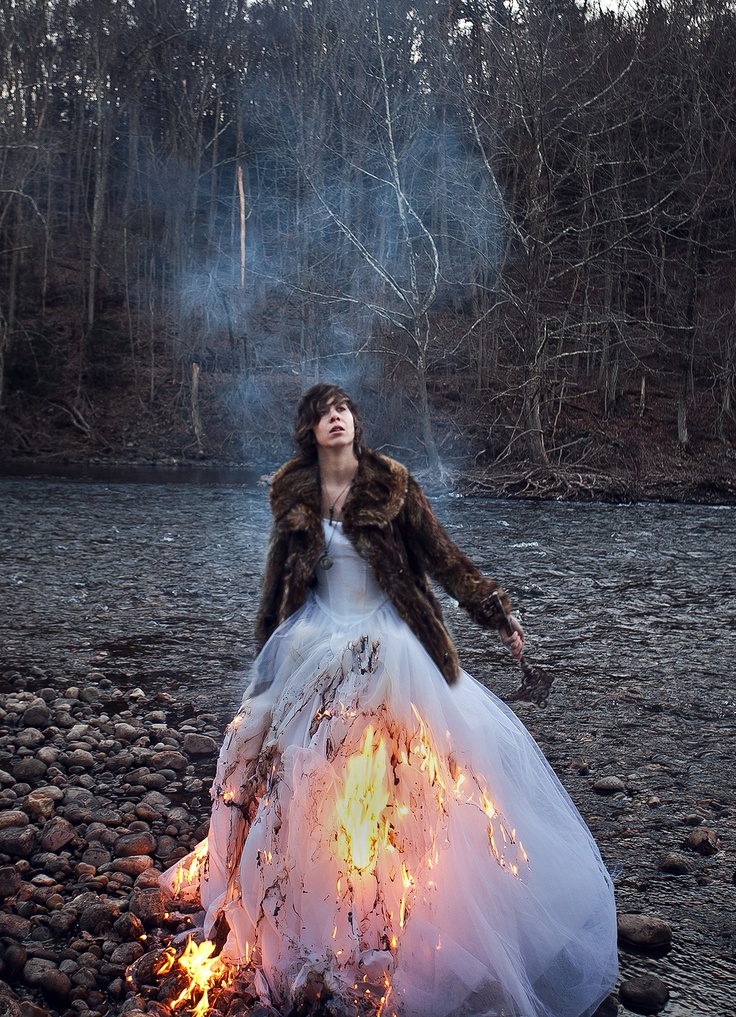 a woman in a long white dress standing next to a fire with her arms outstretched