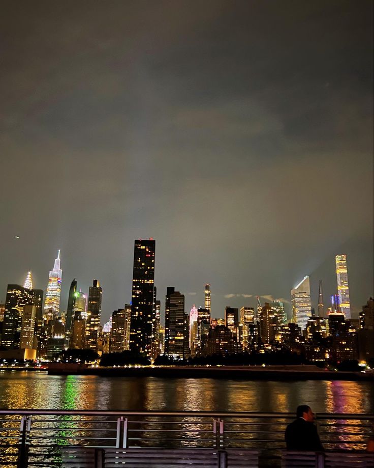 the city skyline is lit up at night with people sitting on benches looking out over the water