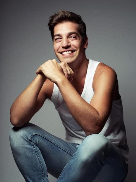 a smiling man sitting on the ground with his arms crossed and legs crossed, wearing blue jeans