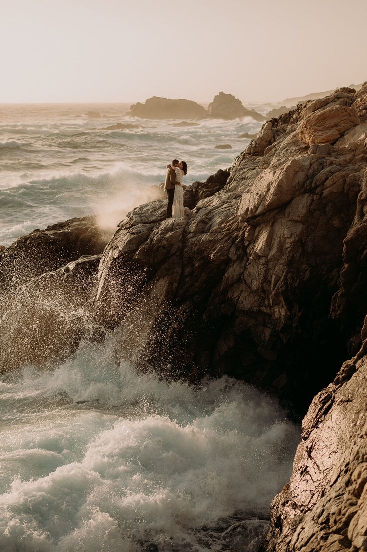 a man standing on top of a rock next to the ocean with waves crashing around him