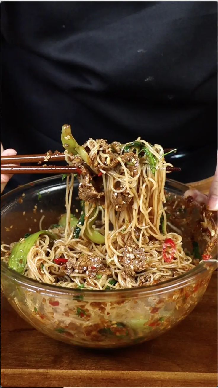 someone holding chopsticks over a bowl of noodles with meat and veggies