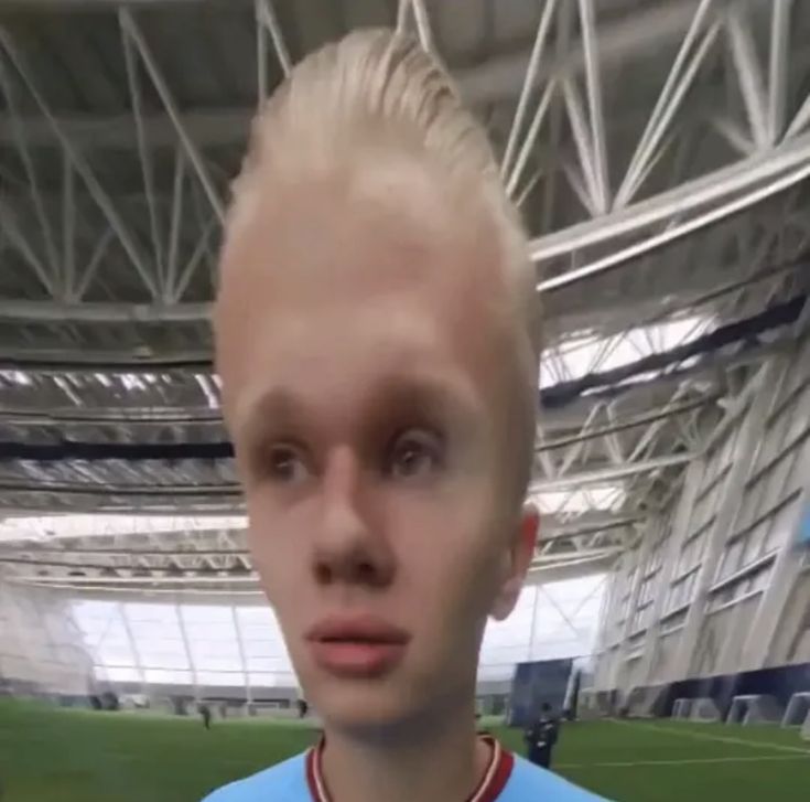 a man with blonde hair standing in front of a soccer field and looking at the camera