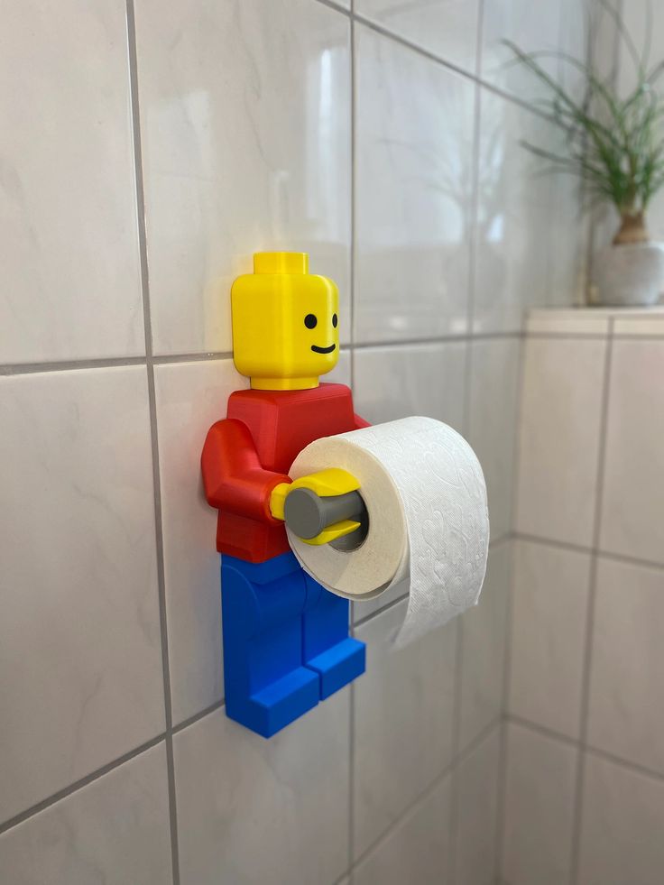 a lego man holding a roll of toilet paper on a bathroom wall with a plant in the corner