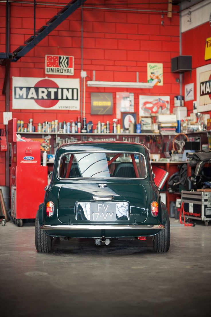 an old green car in a garage with other cars and tools on the wall behind it