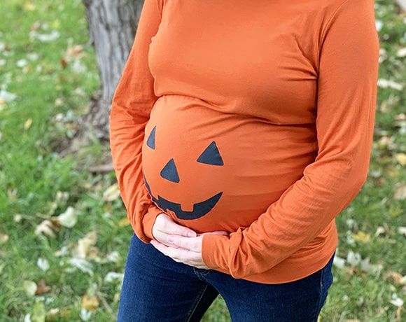 a pregnant woman wearing an orange shirt and pumpkin hat, standing in front of a tree