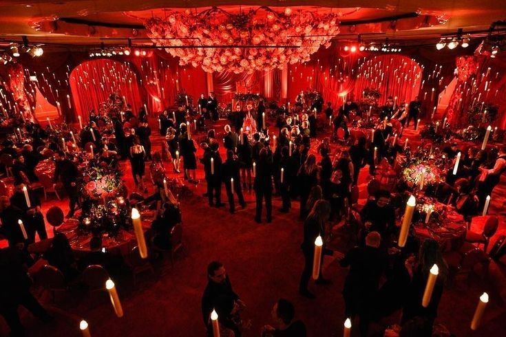 a group of people standing around tables in a room with candles on the floor and red lights hanging from the ceiling