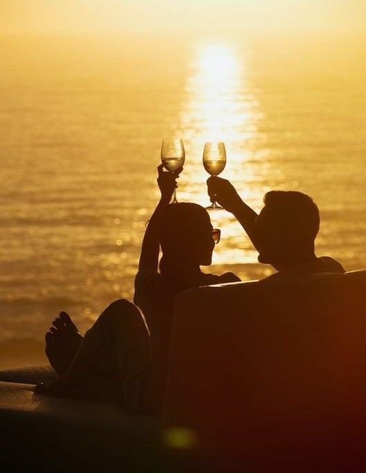 two people toasting with wine glasses in front of the ocean at sunset or sunrise