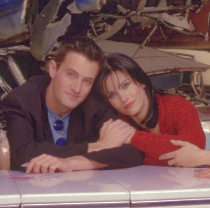 a man and woman leaning on top of a car in front of a pile of junk