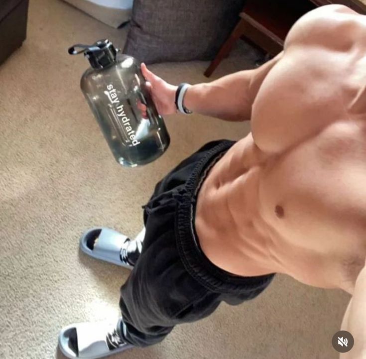 a shirtless man holding a water bottle in his right hand and standing on the floor