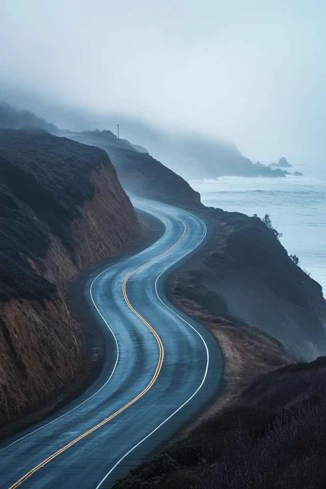 a winding road on the side of a cliff by the ocean in foggy weather