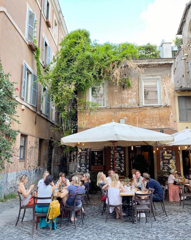 people sitting at tables in an alleyway with umbrellas on the sides and buildings behind them