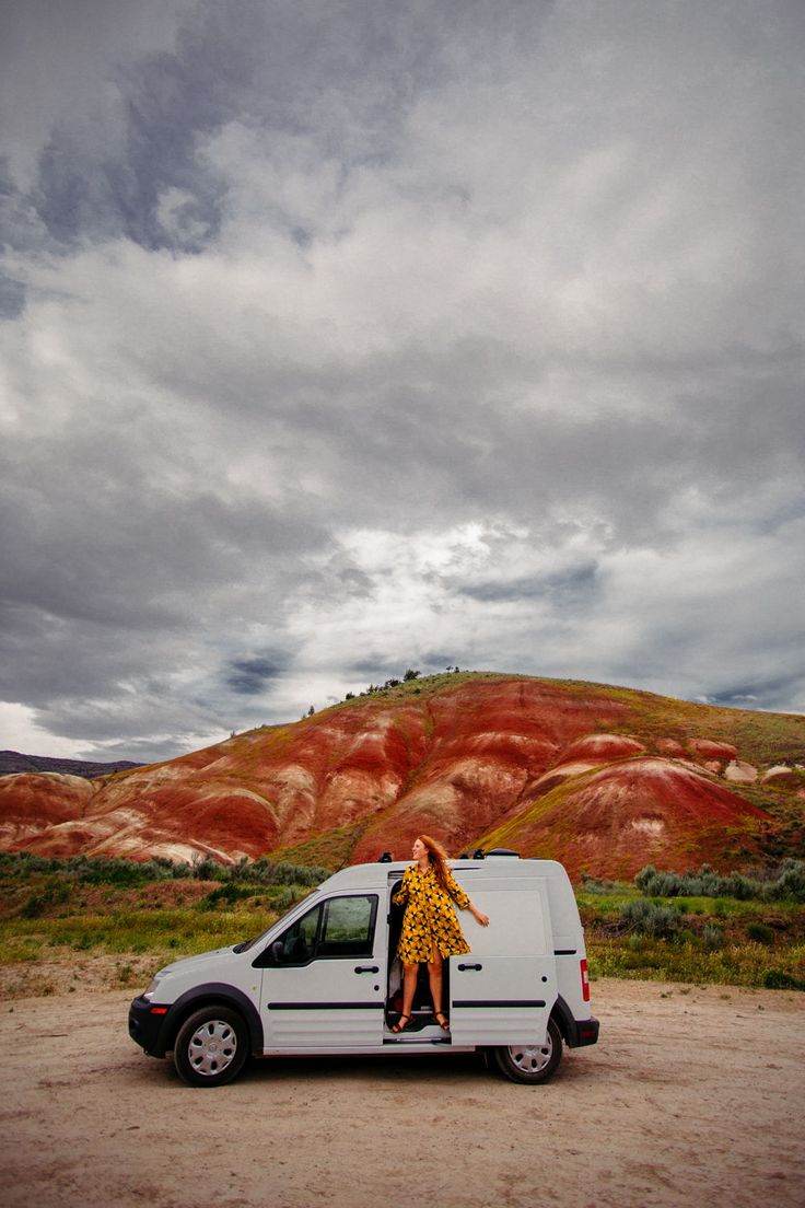 a woman in a yellow dress standing on the back of a white van with mountains behind her