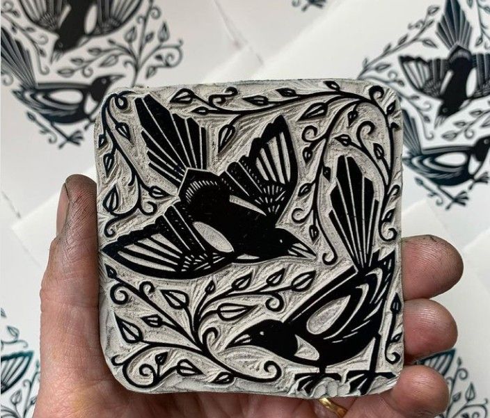 a hand holding up a square tile with a bird on it's side and another design in the background