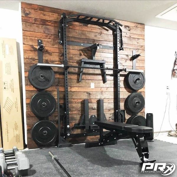 a home gym with two squat racks and several weight plates on the wall, in front of a wood paneled wall