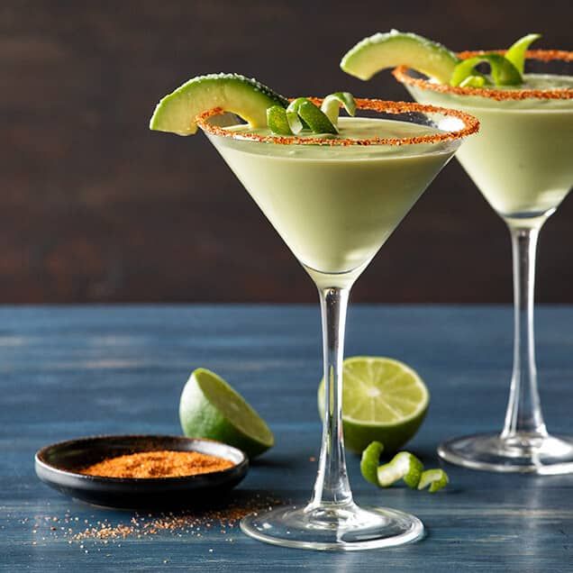 two martini glasses with limes and garnishes on the rim, one filled with an avocado