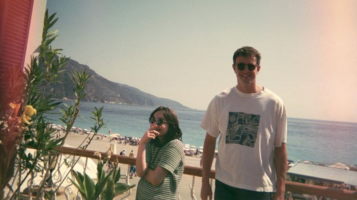 a man and woman standing next to each other on a balcony near the ocean with mountains in the background