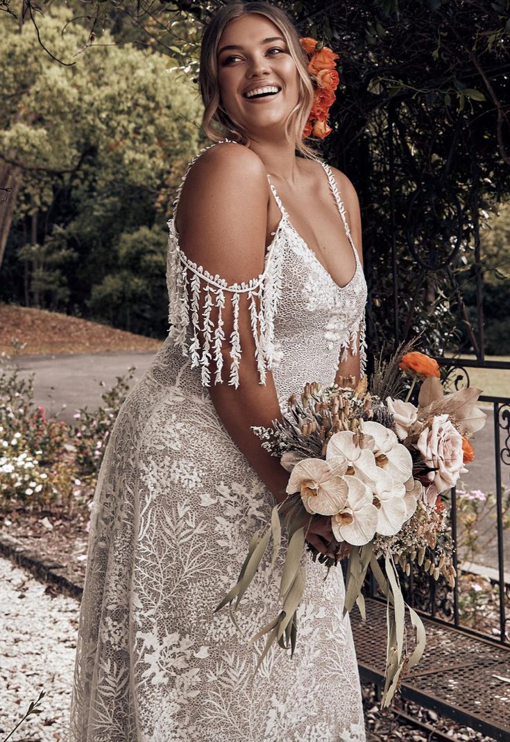 a woman standing in front of a fence holding a bouquet of flowers and wearing a white dress