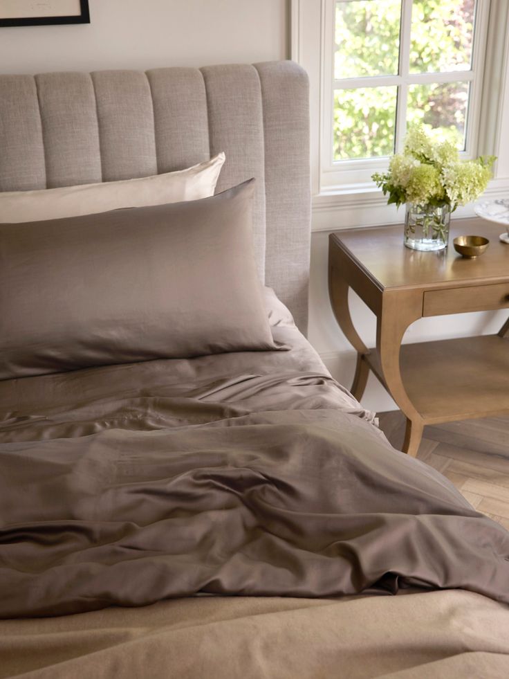 a bed with two pillows on top of it next to a table and chair in front of a window