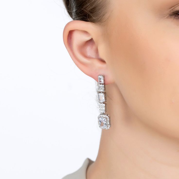 Step into a world of glamour with the Crawford Drop Earrings in Silver White. Exquisitely fashioned  and resplendent with dazzling simulated diamonds, these earrings exude an Art Deco charm that’s both vintage and timeless.  The elegant drop design features five rectangular settings. Each setting is graced with a unique arrangement of gemstones, crowned by a single radiant simulated diamond at the bottom, encircled by a stunning array of baguette-cut stones.   These earrings are designed for bot Luxury Bridal Drop Earrings With Sparkling Stones, Glamorous Brilliant-cut Bridal Earrings For Formal Occasions, Luxury Bridal Drop Earrings With Diamond Accents, Glamorous Brilliant Cut Bridal Earrings For Formal Occasions, Glamorous Brilliant Cut Bridal Earrings For Formal, Luxury Crystal Diamond Drop Earrings, Glamorous Brilliant Cut Earrings For Party, Luxury Crystal Drop Earrings For Anniversary, Glamorous Brilliant Cut Diamond Earrings For Evening