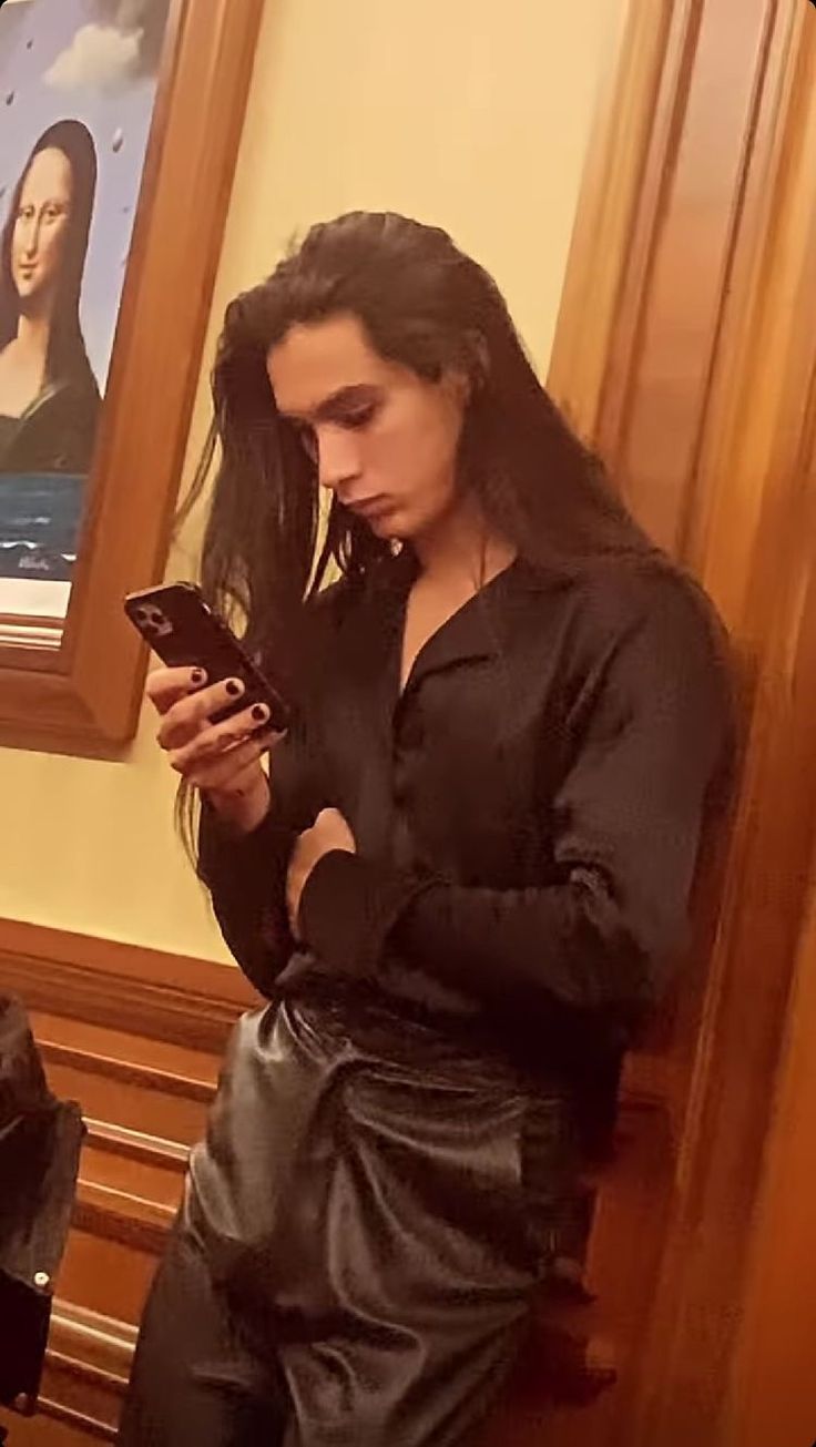 a woman standing in front of a mirror looking at her cell phone while wearing a leather skirt