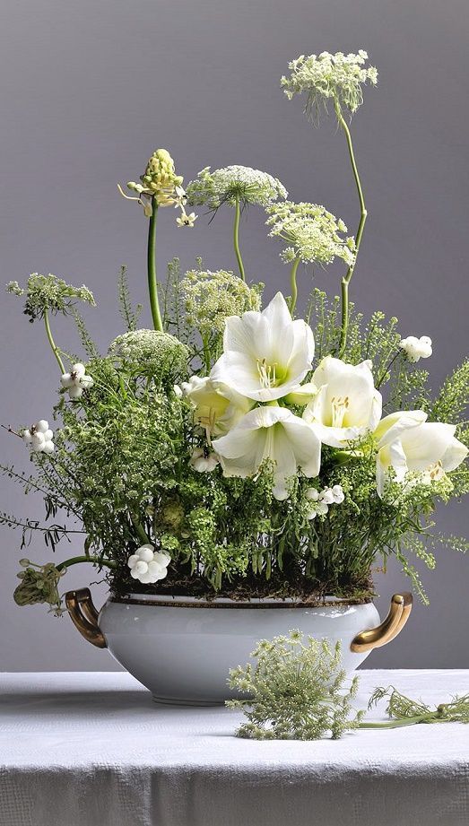 a white flower arrangement in a bowl on a table with greenery and other flowers