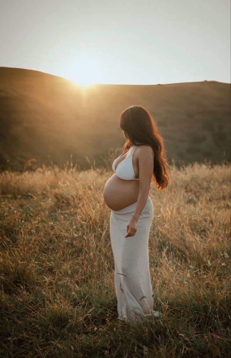 a pregnant woman standing in a field at sunset