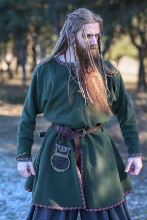 a man with long hair and beard wearing a green outfit standing in the woods holding his hands on his hips