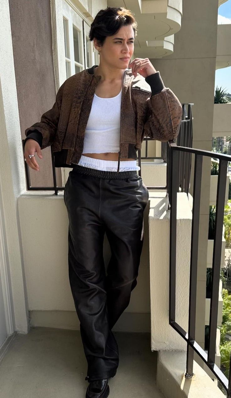 a woman in black leather pants and a white top is standing on a balcony with her hands behind her head