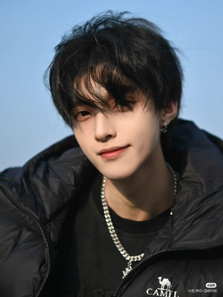 a young man with black hair wearing a black jacket and silver necklace, against a blue sky