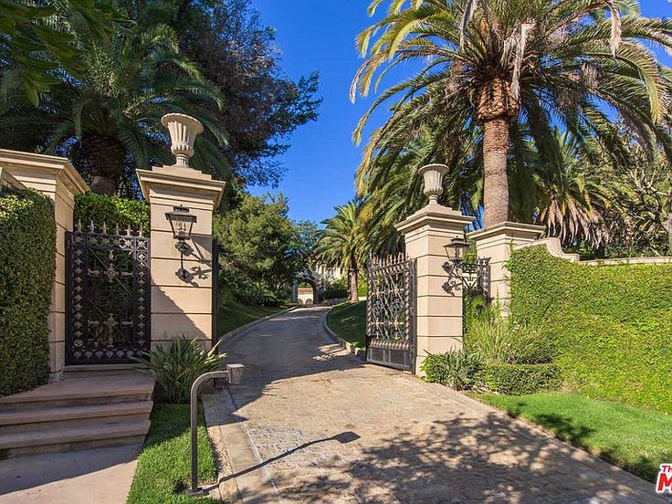 a driveway leading to a gate with palm trees in the background and bushes on either side