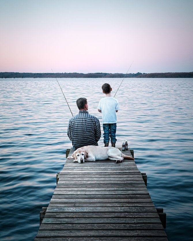 a father and son sitting on a dock with their dog fishing in the water at sunset