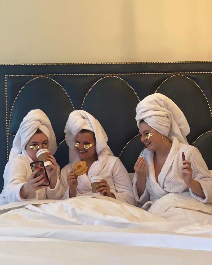 three women in white robes are sitting on a bed and one is eating food while the other looks at her phone