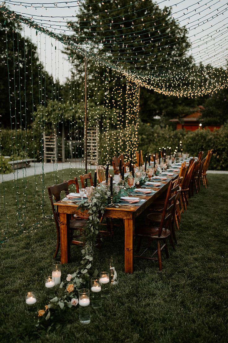 an outdoor dinner table set up with candles and greenery on the grass, surrounded by string lights