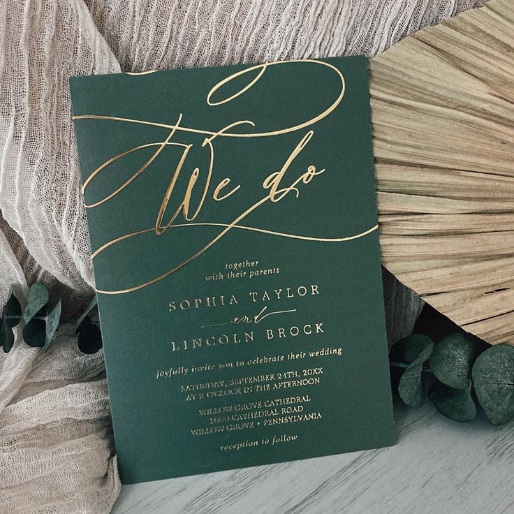 a green and gold wedding card with the word we do written on it, surrounded by greenery