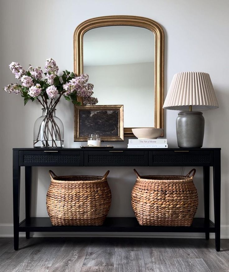 two wicker baskets under a table with a mirror and lamp on it, next to a vase with flowers