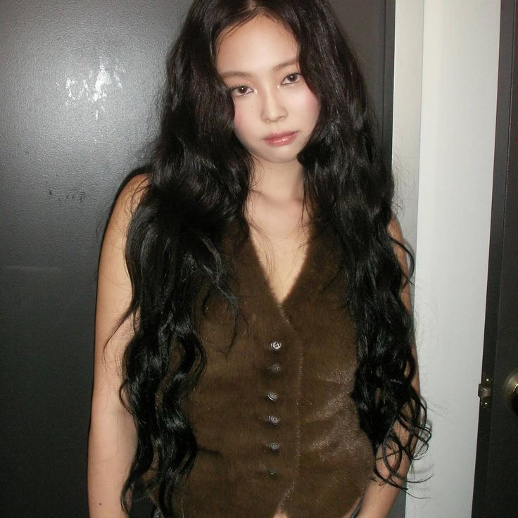 a woman with long black hair standing in front of a door wearing a brown vest