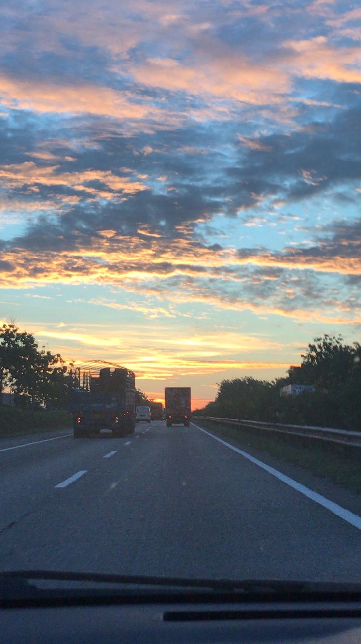 the sun is setting behind two trucks driving down the road with trees on either side