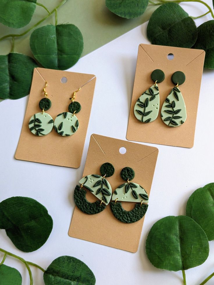 three pairs of green and white earrings sitting on top of a table next to leaves