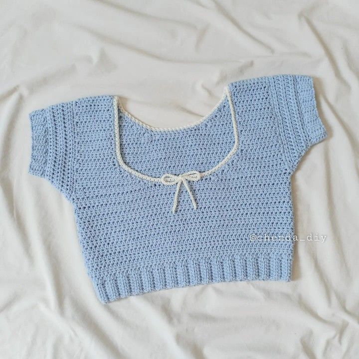 a blue knitted sweater laying on top of a white bed sheet with a knot at the neck