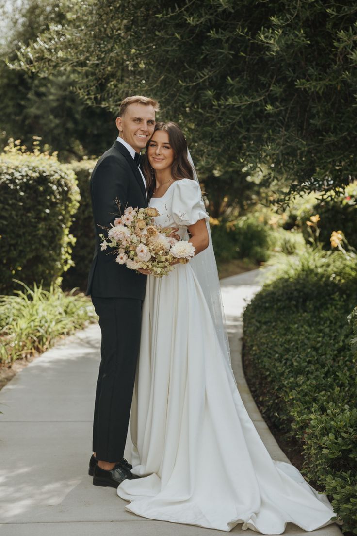 a bride and groom pose for a photo on the sidewalk in front of some bushes